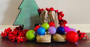 Military boots with Christmas ornaments laid across them and a Christmas tree in the background.