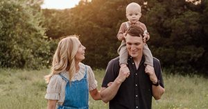 Sydney and her husband walking through a field with their baby on their shoulders.