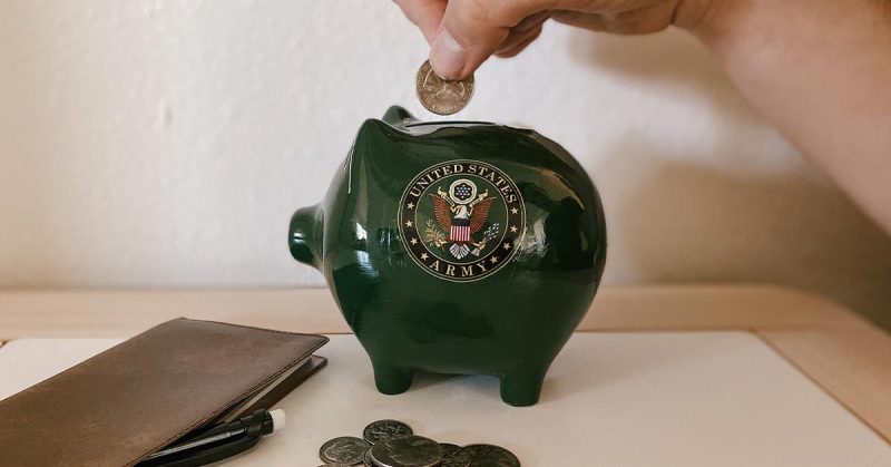 A hand putting money into a green piggy bank with the U.S. Army logo on it.