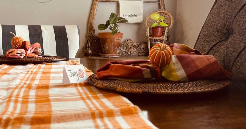A view of a Thanksgiving table is shown, featuring an orange plaid blanket, a pumpkin table-setting and a sign that says “thankful.”