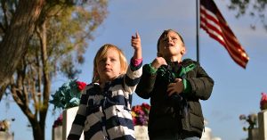 1. A female child and a male child are seen pointing at the camera. In the background, an American flag flies.