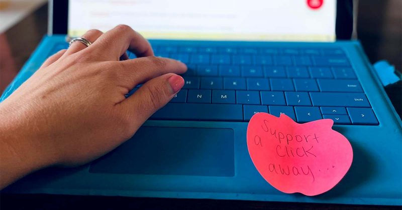 A hand is seen typing on a laptop. A sticky note on the laptop reads “Support a click away…”