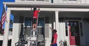 Two people hang string lights on a white house; one on a ladder, one assisting from the ground. American flag visible