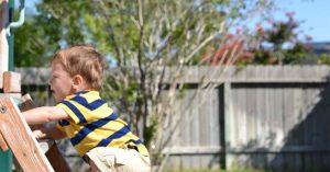 Young boy climbing a wooden playset ladder in a fenced backyard on a sunny day
