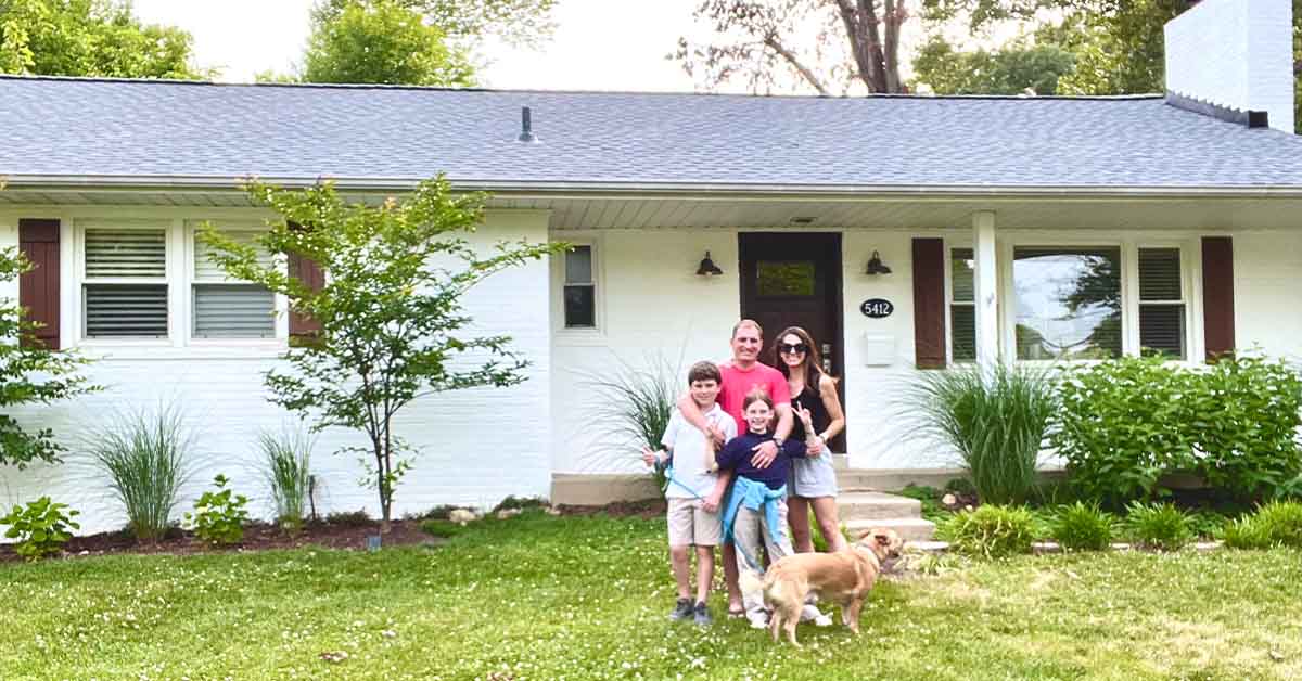 Smiling family of five, including a dog, stands together on the lawn in front of a single-story white house