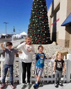 Four children smile in front of a tall, decorated Christmas tree