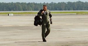 Kristi’s military husband walks across runway beneath wing of large military aircraft, carrying gear bag
