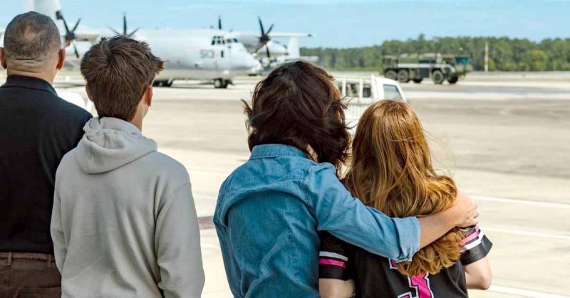Family stands on airfield watching military plane; Kristi hugs daughter
