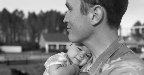 Service member in uniform gently holds a baby against his chest; the baby rests peacefully on his shoulder