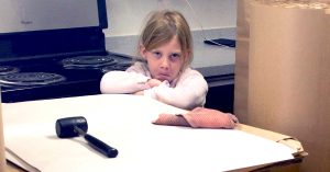 Young girl with crossed arms frowns over a packing station with paper rolls, a mallet and kitchen in background