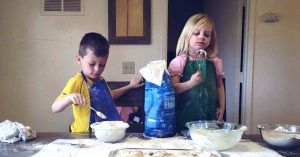 Two kids in aprons baking at a kitchen table with flour, bowls and dough in front of them