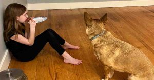 Girl sits on a wood floor eating pizza while a dog sits a few feet away