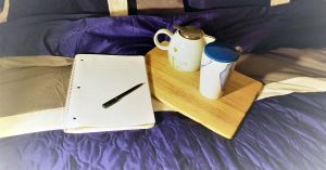 Open notebook and pen beside a wooden tray with a teapot and mug, all placed on a bed
