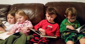 Four kids in pajamas read books together on a brown leather couch, wrapped in blankets