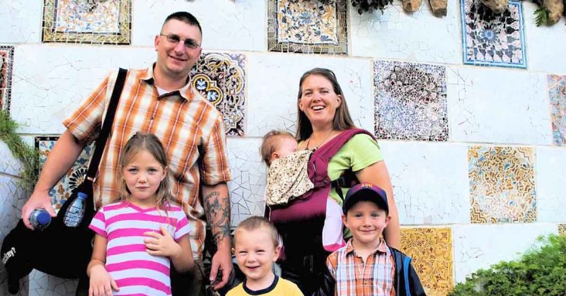 Smiling family of six stands in front of a colorful mosaic tile wall; parents hold baby in carrier.