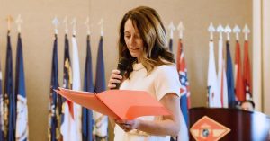 Woman speaks into a microphone while reading from a red folder at a military ceremony.