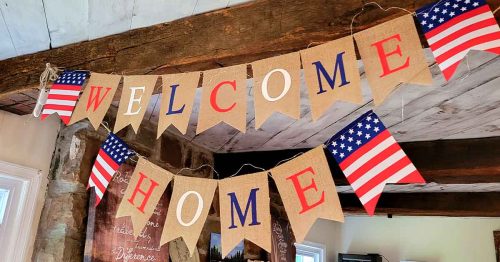 A patriotic “Welcome Home” banner with U.S. flag pennants hangs from wooden beams indoors.