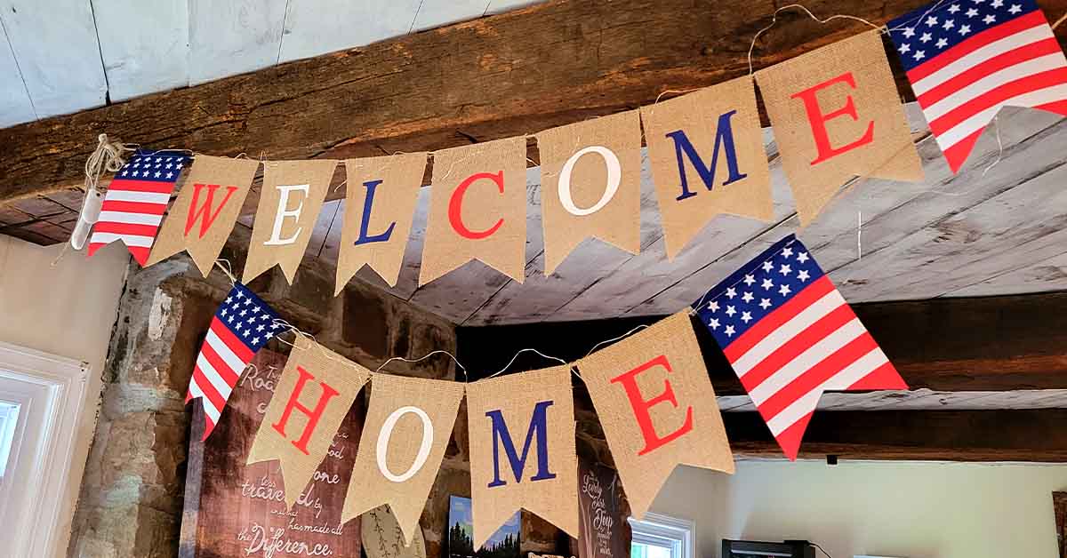 A patriotic “Welcome Home” banner with U.S. flag pennants hangs from wooden beams indoors.