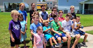 A large group of kids sit and stand together outdoors, eating popsicles and ice cream in a suburban neighborhood.