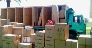 A moving truck is surrounded by cardboard boxes as two movers load large furniture crates.