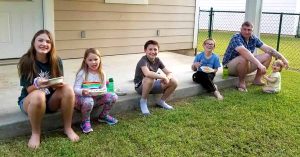 A group sits on a porch step eating food and smiling at the camera.