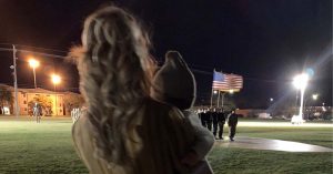 Sydney holding her baby watching a nighttime military ceremony with a raised American flag.