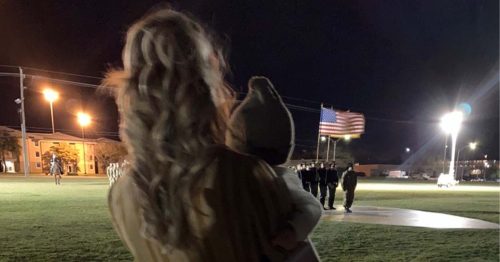 Sydney holding her baby watching a nighttime military ceremony with a raised American flag.