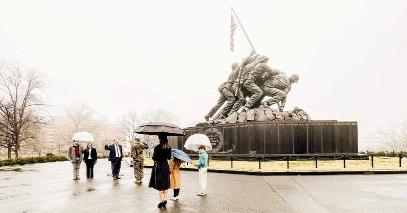 Small group gathers with umbrellas in front of the Marine Corps War Memorial as a military member raises their right hand in oath.