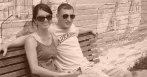 Kristi and her husband wearing sunglasses on a wooden bench against a brick wall.