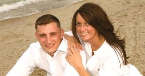 Kristi and her husband in white shirts sitting close together on a sandy beach near the water’s edge.