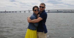 Kristi and his husband wearing sunglasses hugging by the water, with a long bridge stretching across the background.