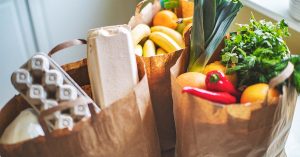 Three paper grocery bags filled with fresh produce, eggs, herbs, bananas and vegetables by a sunny window.