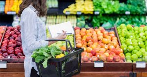 Woman with a shopping list and basket full of produce stands in front of a colorful grocery store apple display.