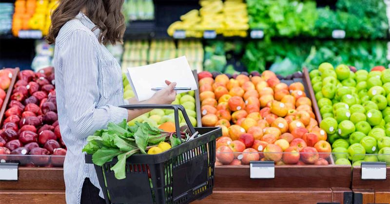 Woman with a shopping list and basket full of produce stands in front of a colorful grocery store apple display.