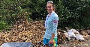 Kristi outdoors holding a wheelbarrow, standing near a pile of yard debris and greenery