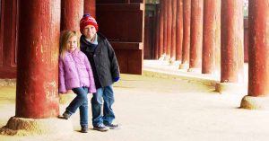 Kristi’s children, in jackets, stand beside red columns in a walkway, smiling at the camera