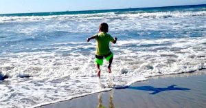 Kristi’s child, in a green outfit, runs and jumps into ocean waves on a sandy beach