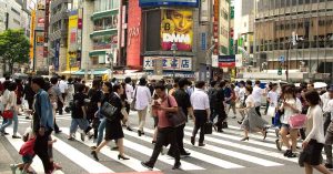 Crowded city crosswalk filled with pedestrians, surrounded by tall buildings and large digital billboards