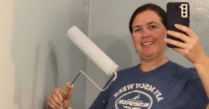 Lizann takes a mirror selfie while painting a wall with a roller, wearing a casual T-shirt.
