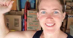 Lizann smiles and flexes her arm in front of stacked moving boxes in a garage or storage space.