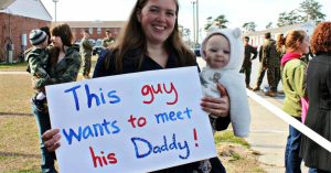 A woman holds a baby and a sign reading, “This guy wants to meet his Daddy!” with people in military uniforms in the background