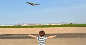 A young child stands with arms outstretched facing a low-flying aircraft over an open desert landscape