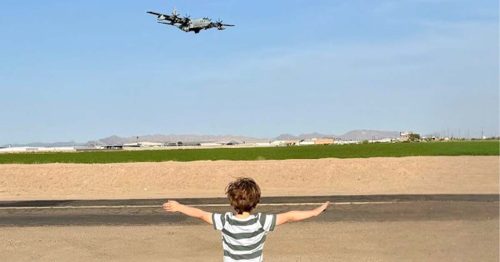A young child stands with arms outstretched facing a low-flying aircraft over an open desert landscape