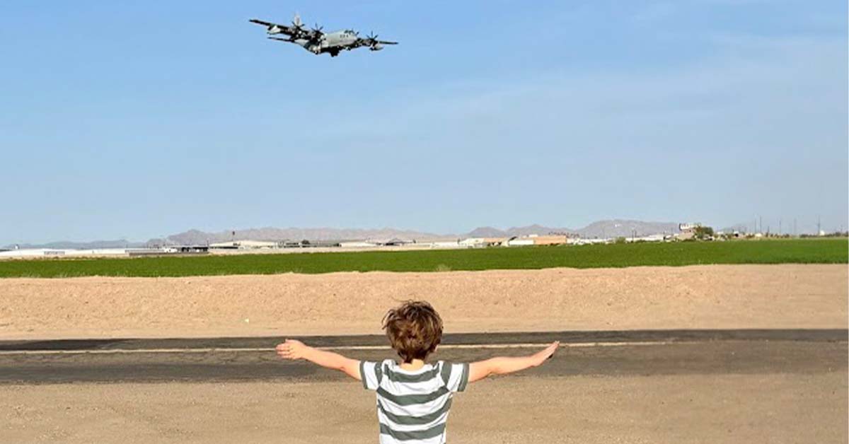 A young child stands with arms outstretched facing a low-flying aircraft over an open desert landscape
