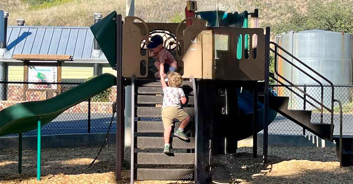 Two young children play on a playground structure, one helping the other up the stairs near a slide