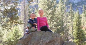 Lizann’s children sit on a large boulder in a forested mountain area, smiling, with tall pine trees and rocky cliffs behind them