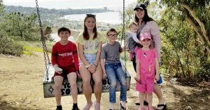 Lizann and her children stand and sit on a large outdoor swing overlooking a coastal city and water below