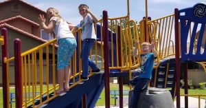 Lizann’s children play on a colorful playground structure, climbing stairs and waving