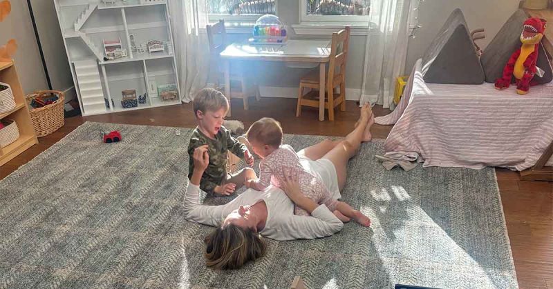 A sunlit playroom where Sydney lies on a rug while her young child and a baby climb and play, surrounded by toys and furniture