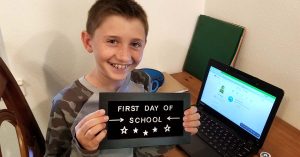 Lizann’s smiling son holding a "First Day of School" sign while seated at a desk with a laptop.